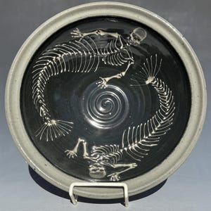 May include: A ceramic bowl with a dark interior and a light gray rim. The bowl features a white skeletal design of two creatures in a circular pattern. The bowl is displayed on a white metal stand.