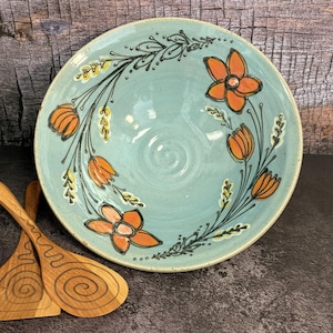 May include: A round, light blue ceramic bowl with a speckled finish, featuring hand-painted orange flowers and black leafy stems. Two light-colored wooden serving spoons with dark, spiral-patterned engravings are positioned next to the bowl.