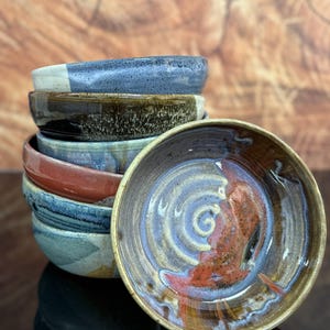 May include: A stack of six round ceramic bowls with varied glaze finishes. The top bowl features a glossy interior with swirling patterns of brown, blue, and cream. Other bowls in the stack display mottled textures and colors including blue, brown, and red.