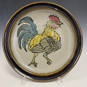 May include: A ceramic plate featuring a hand-painted rooster design. The rooster is yellow with a red comb and wattle, and blue-green tail feathers. The plate has a speckled beige center and a dark brown rim, with a black outer edge.