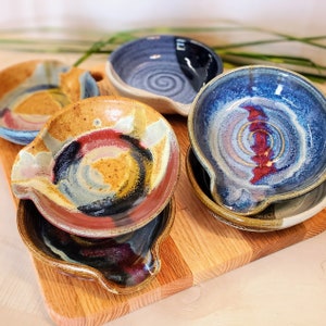 May include: A collection of handmade ceramic spoon rests in various colors and designs. The spoon rests feature a circular shape with a handle rest, and are decorated with swirls of blue, red, yellow, and brown. They are arranged on a wooden cutting board.