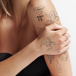 May include: Black temporary tattoos with wedding-themed designs, including a champagne bottle, cake, rings, and the phrase "Happily Ever After." The tattoos are applied on the arm and shoulder.