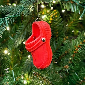 Puede incluir: Adorno navideño de zueco rojo brillante colgando de un árbol de Navidad verde. El adorno es una pequeña réplica de un zapato de zueco, con un detalle de botón plateado. El árbol está decorado con luces blancas.