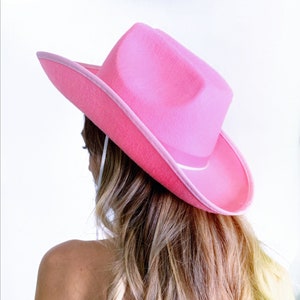 K&ouml;nnte beinhalten: Ein rosa Cowboyhut mit wei&szlig;em Band. Der Hut wird von einer Person mit langen blonden Haaren getragen.