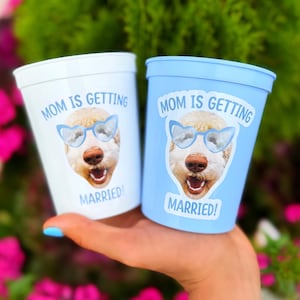 Peut inclure: Deux gobelets en plastique, l'un blanc et l'autre bleu clair, arborant chacun l'image d'un chien portant des lunettes de soleil bleues en forme de c&oelig;ur avec le texte "MOM IS GETTING MARRIED!" imprim&eacute; en bleu au-dessus et en dessous.