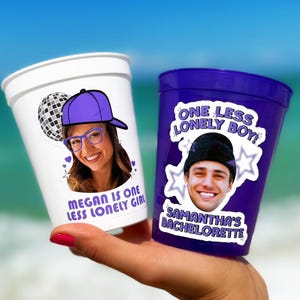 May include: A white plastic cup featuring a disco ball, a person wearing a purple cap and glasses, and the text "MEGAN IS ONE LESS LONELY GIRL" alongside a purple plastic cup with a person in a black beanie and the text "ONE LESS LONELY BOY" and "SAMANTHA'S BACHELORETTE".