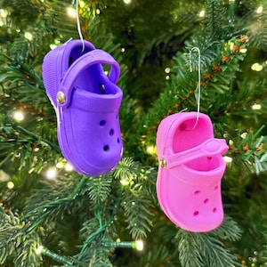 Puede incluir: Dos adornos de zapatos Crocs en miniatura, uno morado y otro rosa, cuelgan de un árbol de Navidad. Los adornos tienen un diseño de zueco clásico con una correa y agujeros de ventilación. Cada zapato está suspendido por un gancho de alambre plateado.