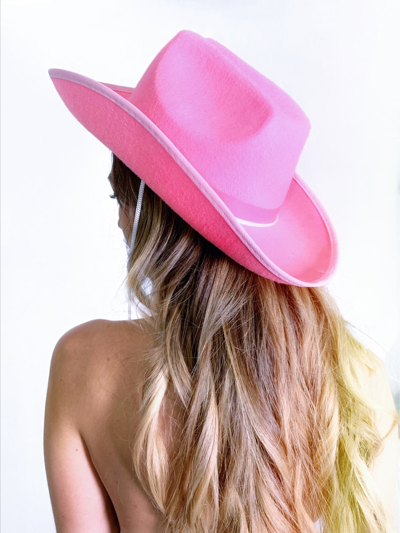 K&ouml;nnte beinhalten: Ein rosa Cowboyhut mit wei&szlig;em Band. Der Hut wird von einer Person mit langen blonden Haaren getragen.