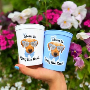 Peut inclure: Un gobelet en plastique blanc et un gobelet en plastique bleu clair, chacun orn&eacute; d'un dessin de chien marron portant des lunettes bleu clair et un n&oelig;ud papillon assorti, avec le texte "Mom is Tying the Knot" imprim&eacute; en noir autour du chien.