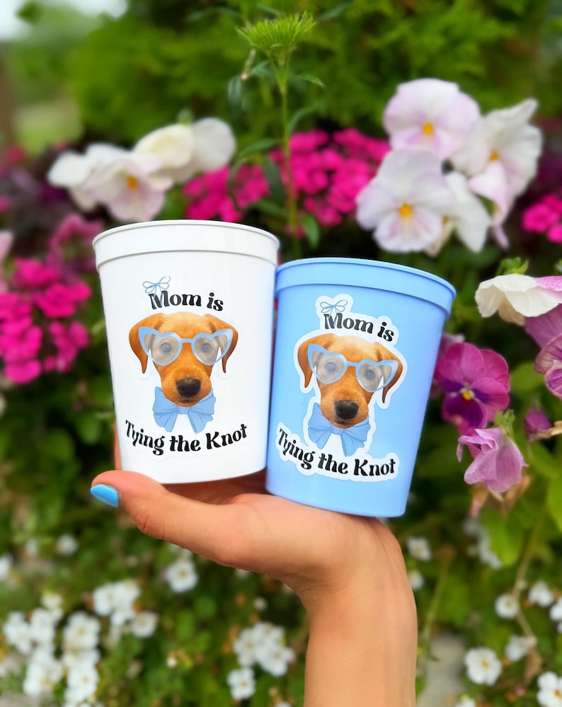 Peut inclure: Un gobelet en plastique blanc et un gobelet en plastique bleu clair, chacun orn&eacute; d'un dessin de chien marron portant des lunettes bleu clair et un n&oelig;ud papillon assorti, avec le texte "Mom is Tying the Knot" imprim&eacute; en noir autour du chien.