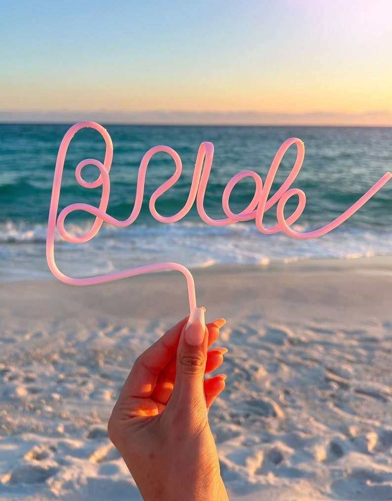 May include: Pink plastic straw bent into the word "Bride" against a background of a beach and ocean at sunset.