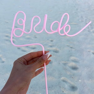 May include: Pink plastic straw bent into the word "Bride" for a wedding celebration.