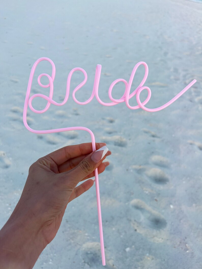 May include: Pink plastic straw bent into the word "Bride" for a wedding celebration.