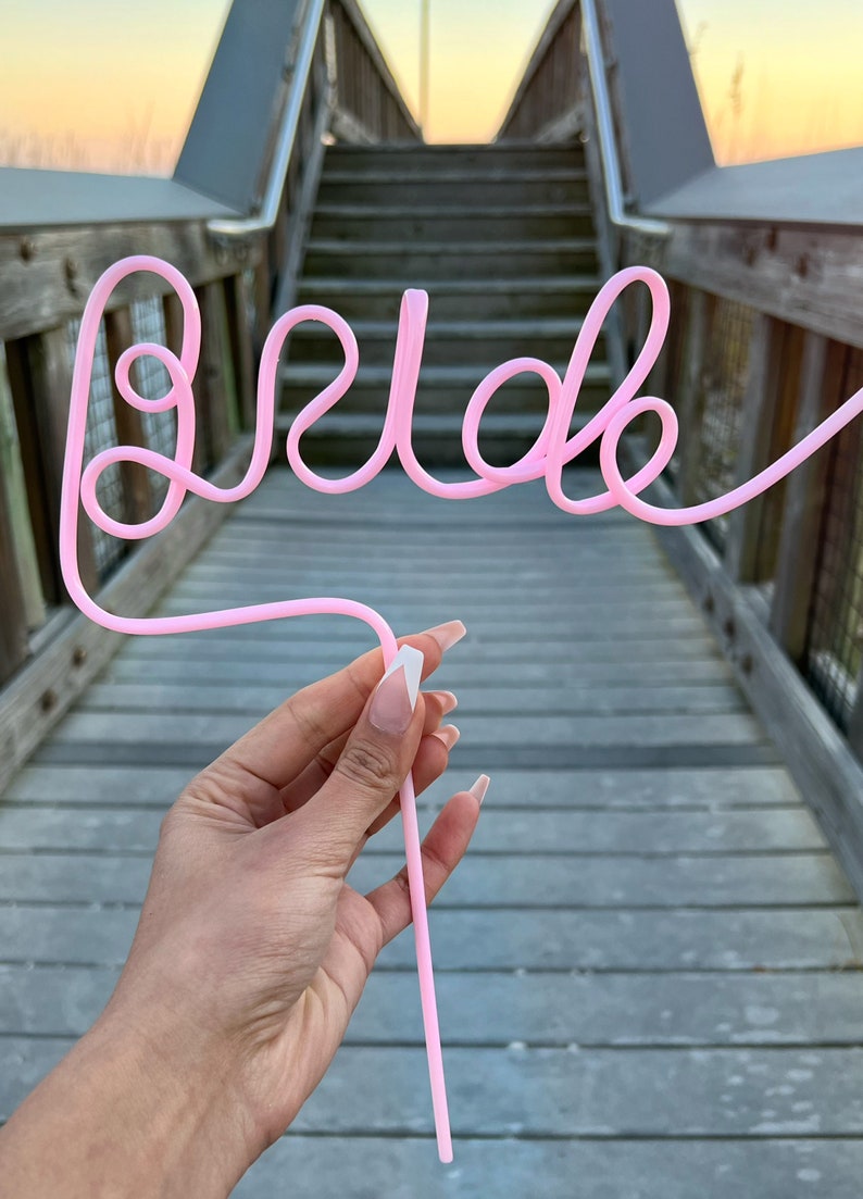May include: Pink plastic straw bent into the word "Bride" with a white tip. The straw is held by a hand with pink nail polish.