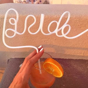May include: A white plastic straw bent into the word "bride" with an orange slice in a glass of pink cocktail.