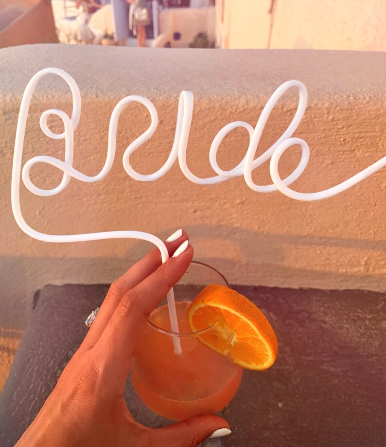 May include: A white plastic straw bent into the word "bride" with an orange slice in a glass of pink cocktail.