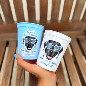 Peut inclure: Un gobelet en plastique bleu clair avec le texte Emma's Last toast on the coast Cape May 2026 et un gobelet blanc avec le texte Mom is getting married. Les deux pr&eacute;sentent l'image d'un chien noir portant des lunettes de soleil en forme de c&oelig;ur.