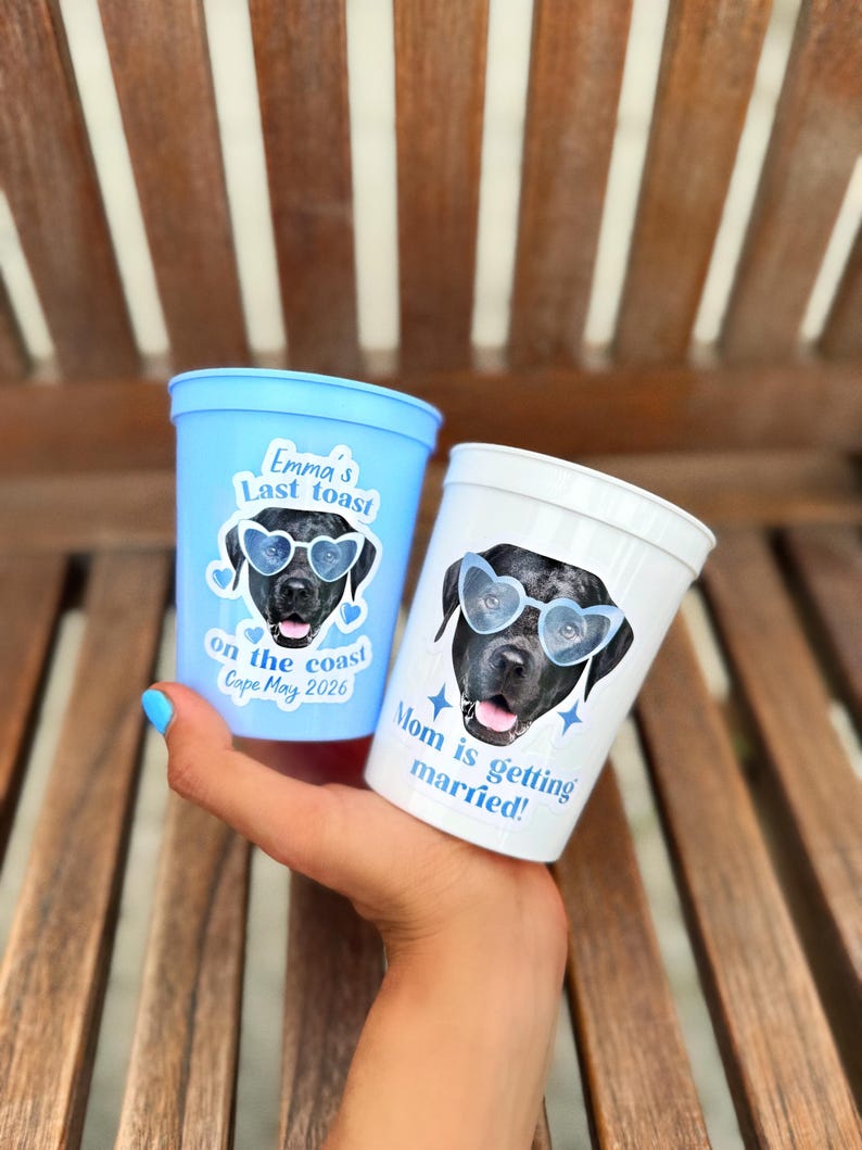 Peut inclure: Un gobelet en plastique bleu clair avec le texte Emma's Last toast on the coast Cape May 2026 et un gobelet blanc avec le texte Mom is getting married. Les deux pr&eacute;sentent l'image d'un chien noir portant des lunettes de soleil en forme de c&oelig;ur.