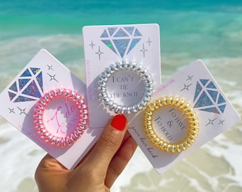 May include: Three colorful hair ties with sayings on white cards. The hair ties are silver, pink, and gold. The sayings are "I Can't Tie The Knot", "I Do Crew", and "To Have & To Hold".