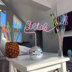May include: A white table with a silver disco ball and a pineapple with colorful straws. The straws are arranged to spell "bride".