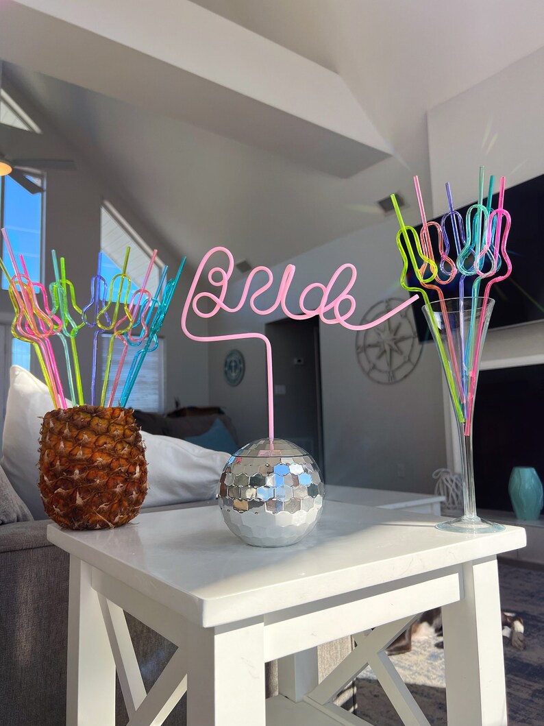 May include: A white table with a silver disco ball and a pineapple with colorful straws. The straws are arranged to spell "bride".
