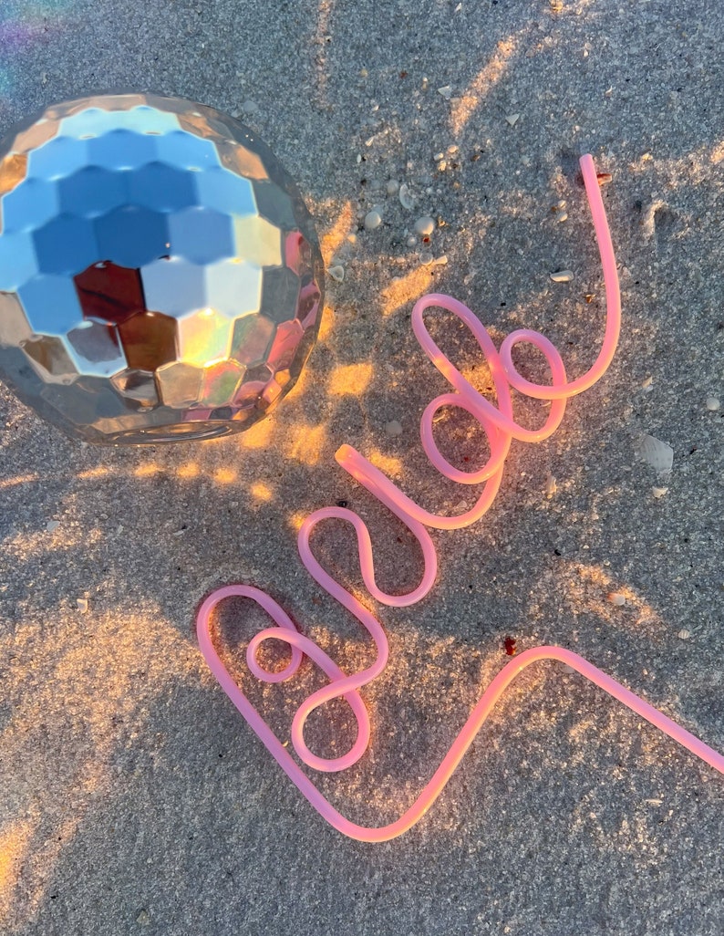 May include: A pink plastic straw bent into the word "bride" on a sandy beach with a silver disco ball in the background.