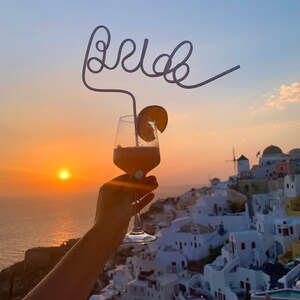 May include: A glass of pink cocktail with a fruit garnish and a straw shaped like the word "bride" in front of a sunset over a white town.