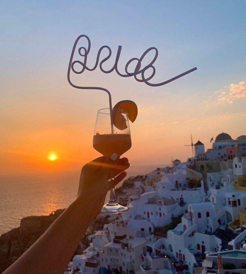 May include: A glass of pink cocktail with a fruit garnish and a straw shaped like the word "bride" in front of a sunset over a white town.