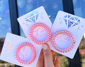 May include: Three pink spiral hair ties with silver accents, each on a white card with a diamond design and the text "I Do Crew". The cards are held in a hand.