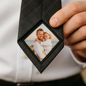 Könnte beinhalten: Eine schwarze Krawatte mit einem rautenförmigen Fotorahmen. Der Rahmen zeigt ein Farbfoto eines Mannes und eines Kindes, die sich umarmen. Die Krawatte hat ein dezentes Karomuster und wird von einer Person gehalten.