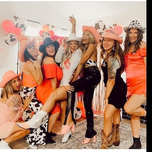 K&ouml;nnte beinhalten: Eine Gruppe von Freundinnen in rosa und wei&szlig;en Cowboyh&uuml;ten und verschiedenen Outfits posiert f&uuml;r ein Foto. Die Frauen l&auml;cheln und lachen, und das Foto f&auml;ngt eine lustige und festliche Atmosph&auml;re ein.
