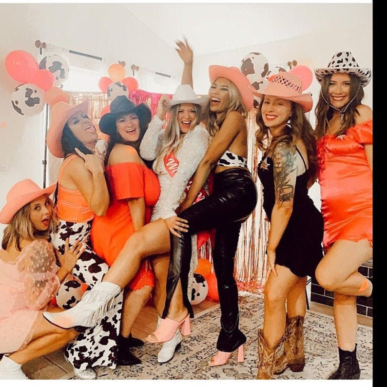K&ouml;nnte beinhalten: Eine Gruppe von Freundinnen in rosa und wei&szlig;en Cowboyh&uuml;ten und verschiedenen Outfits posiert f&uuml;r ein Foto. Die Frauen l&auml;cheln und lachen, und das Foto f&auml;ngt eine lustige und festliche Atmosph&auml;re ein.