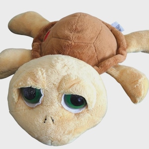 May include: A soft toy turtle featuring a brown shell and tan limbs. The turtle's head is a lighter tan with large, green-eyed, sad-looking eyes. The toy is designed to be soft and cuddly, suitable for children.