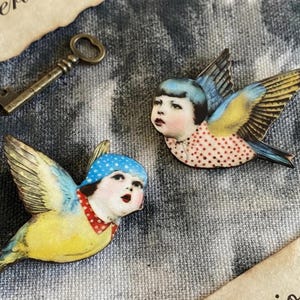 May include: Two whimsical bird-shaped brooches with human faces, featuring blue and yellow wings, red collars, and polka-dotted shirts. A vintage key and a mushroom are also present, suggesting a fairy tale theme.