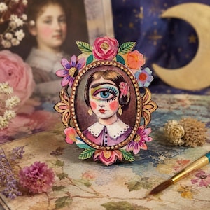 May include: An oval wooden plaque featuring a painted portrait of a person with a single large blue eye in the center of their forehead. The frame is decorated with colorful painted flowers and leaves in shades of pink, orange, and purple.