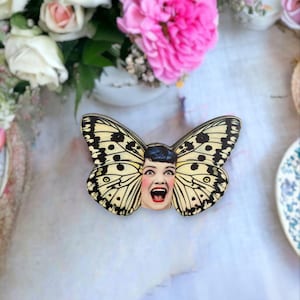 May include: A whimsical butterfly-shaped brooch with a vintage-style image of a woman's face. The butterfly wings are cream-colored with black spots and lines. The woman's face has red lips and a surprised expression. The brooch is a unique accessory.