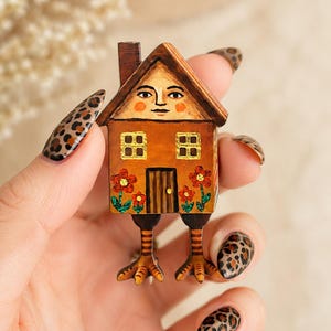 May include: A brown house-shaped brooch with a painted face in the attic window, two square windows, a central door, and red flowers on the facade. The structure is supported by two striped bird-like legs with three-toed feet at the base.