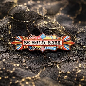 May include: A rectangular enamel pin with a decorative scalloped edge featuring the text BE BOLD, BABE. in black capital letters. The design includes a central white banner surrounded by colorful patterns of red, blue, yellow, and black checkered motifs.