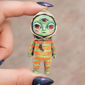 May include: A flat, die-cut wooden pin featuring a three-eyed green creature with large red eyes, wearing a hooded garment with horizontal orange and green stripes and a gold crescent moon emblem centered on the chest area of the figure.