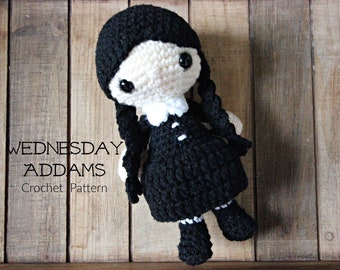 May include: Crochet pattern for a Wednesday Addams doll. The doll is black and white with black hair in braids. The doll is lying on a wooden surface.
