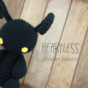 May include: A black crochet plush toy with yellow eyes and a bunny-like appearance. The text "HEARTLESS Crochet Pattern" is displayed on a wooden surface.