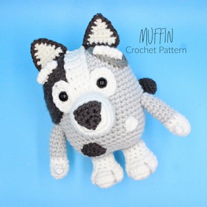 May include: A crocheted stuffed animal dog with grey and white fur, black eyes, and a black nose. The dog is sitting on a blue background and the text "MUFFIN Crochet Pattern" is written above the dog.