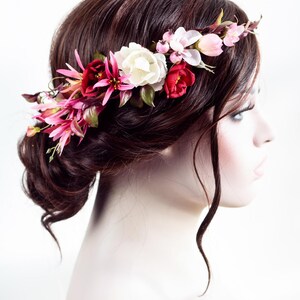 May include: A floral hair crown featuring an arrangement of artificial flowers in shades of deep red, bright pink, and cream white, accented with small green leaves and tiny pink berries, positioned across the top of dark brown hair.