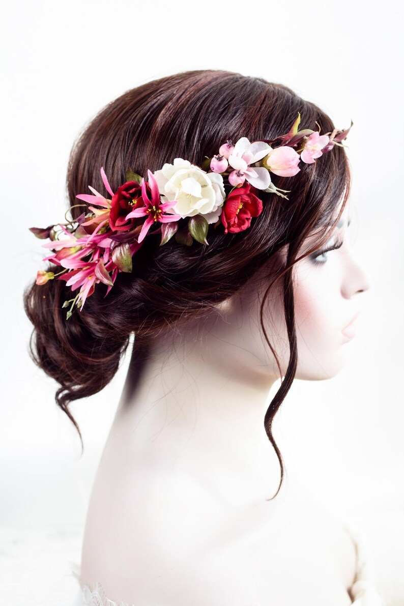 May include: A floral hair crown featuring an arrangement of artificial flowers in shades of deep red, bright pink, and cream white, accented with small green leaves and tiny pink berries, positioned across the top of dark brown hair.