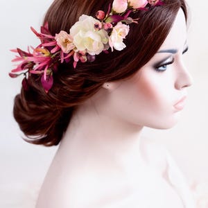 May include: A floral hair crown featuring an arrangement of cream-colored roses, small pink buds, and deep magenta-colored foliage. The accessory is positioned across the top of the head, showcasing a mix of soft pastel and vibrant berry tones.