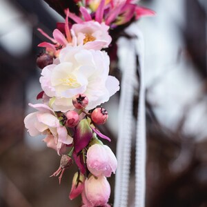 May include: A floral crown featuring a mix of white and pink blossoms, deep magenta buds, and small dark berries. The arrangement is accented with thin white ribbons that hang down, creating a delicate and layered botanical accessory.