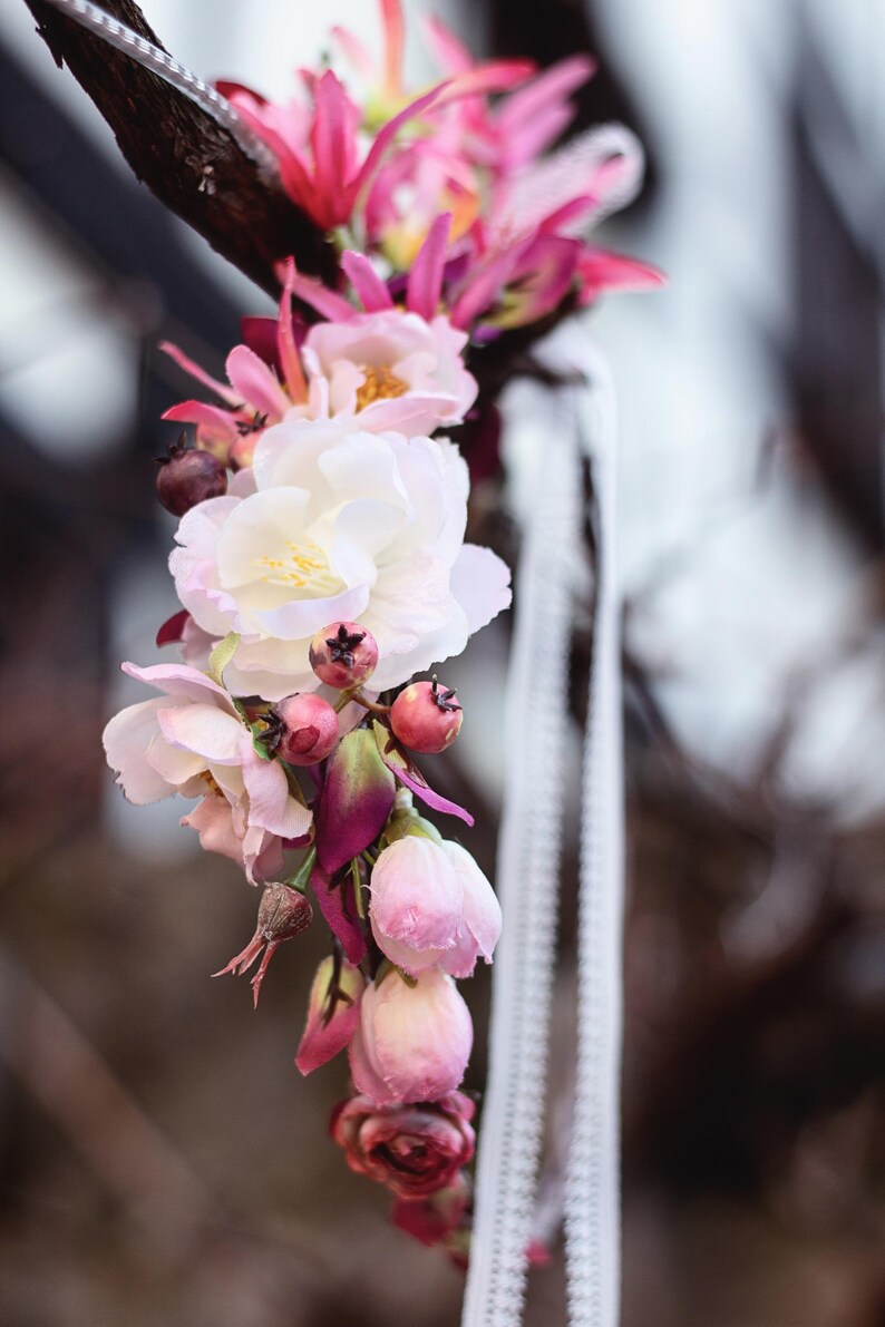 May include: A floral crown featuring a mix of white and pink blossoms, deep magenta buds, and small dark berries. The arrangement is accented with thin white ribbons that hang down, creating a delicate and layered botanical accessory.