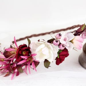 May include: A floral crown featuring a braided brown base adorned with deep burgundy, bright fuchsia, and soft white flowers, accented with small purple berries and dark green leaves, arranged in a cluster along the front of the circular band.