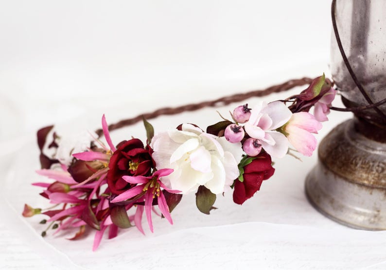 May include: A floral crown featuring a braided brown base adorned with deep burgundy, bright fuchsia, and soft white flowers, accented with small purple berries and dark green leaves, arranged in a cluster along the front of the circular band.
