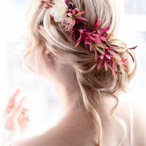 May include: A floral hair accessory featuring a collection of white, light pink, and deep magenta flowers and foliage. The arrangement is tucked into a blonde updo hairstyle, with delicate petals and slender leaves extending along the side.